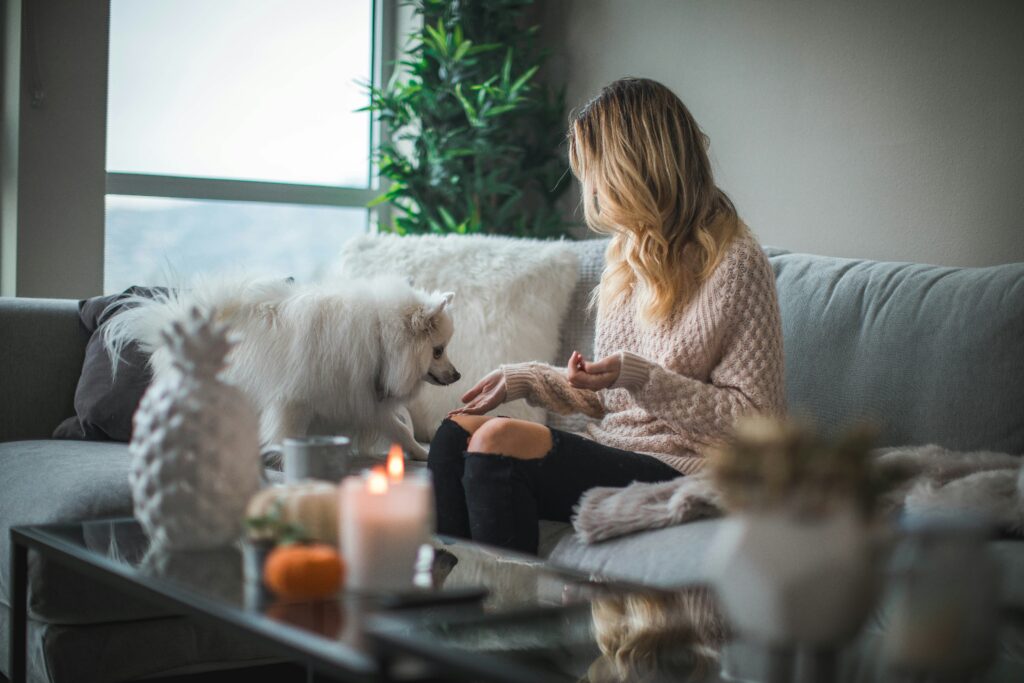 Woman sitting on a sofa holding food for her dog, who is attentively looking at it.