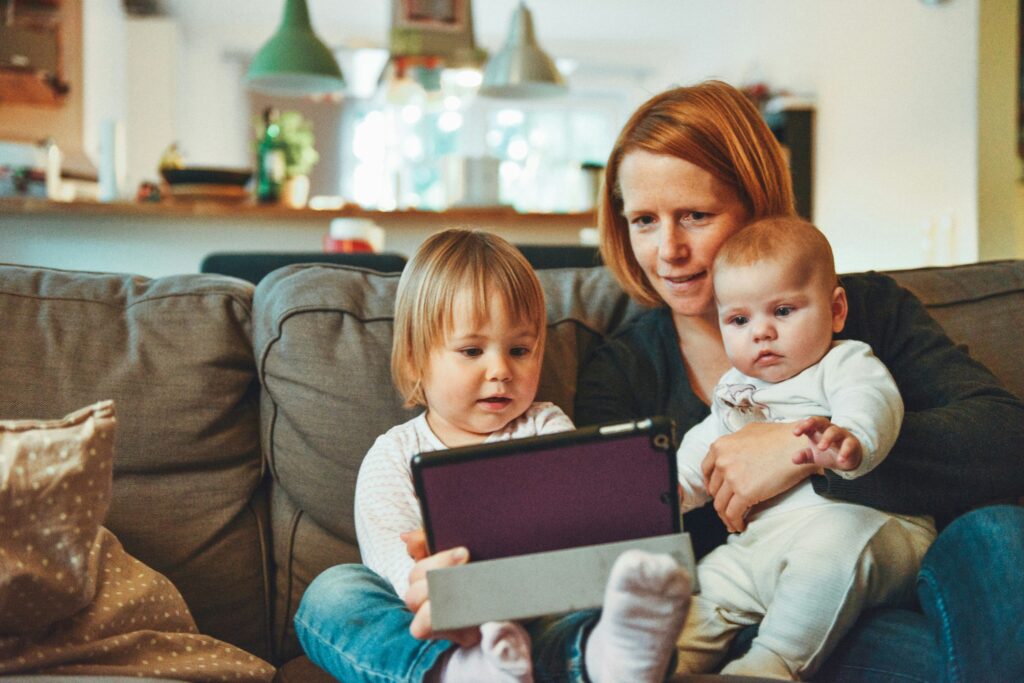 Woman sitting on a sofa holding a baby while watching a tablet with two young children.