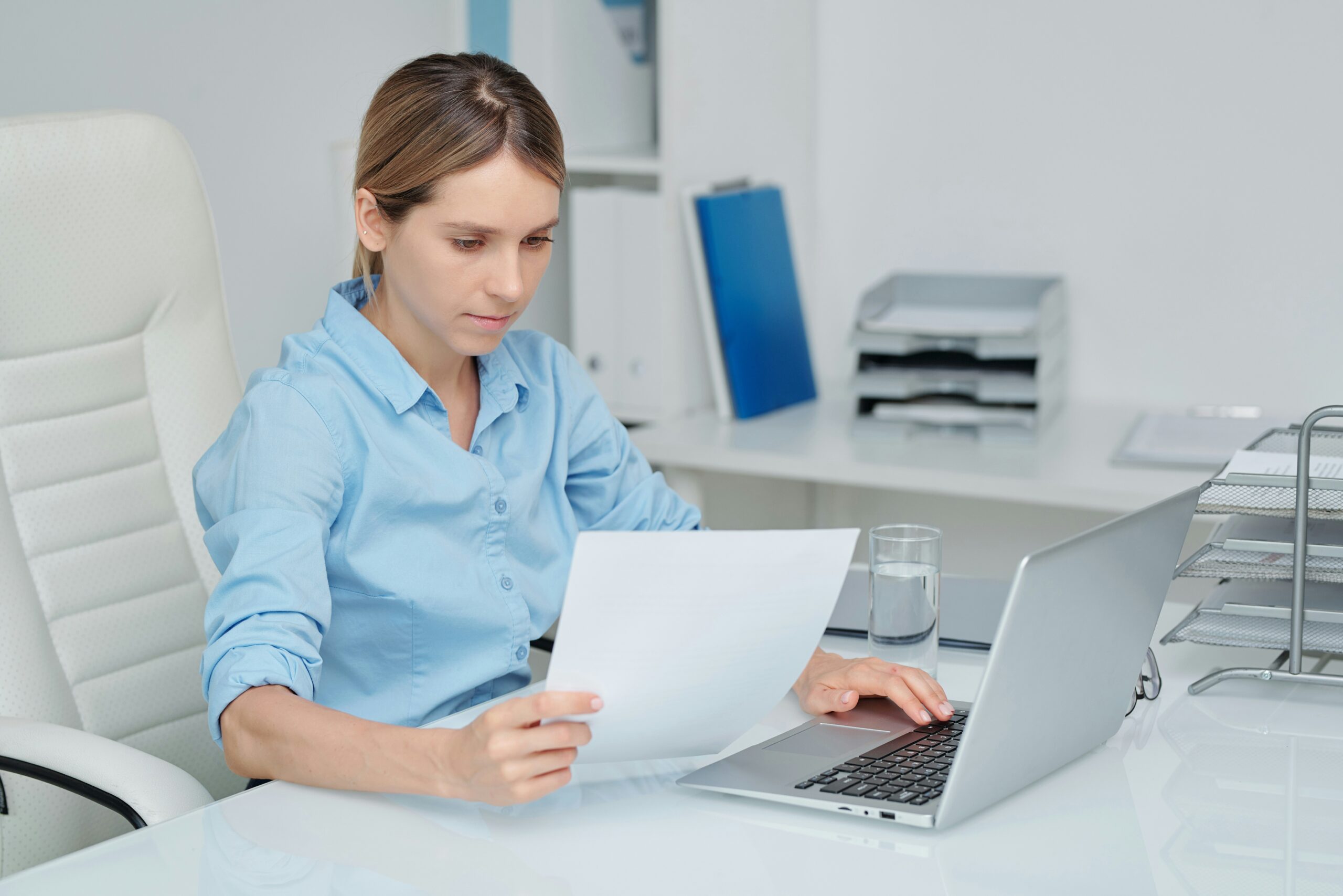 Young professional woman reading a document and reviewing applications on a computer while sitting at a desk.